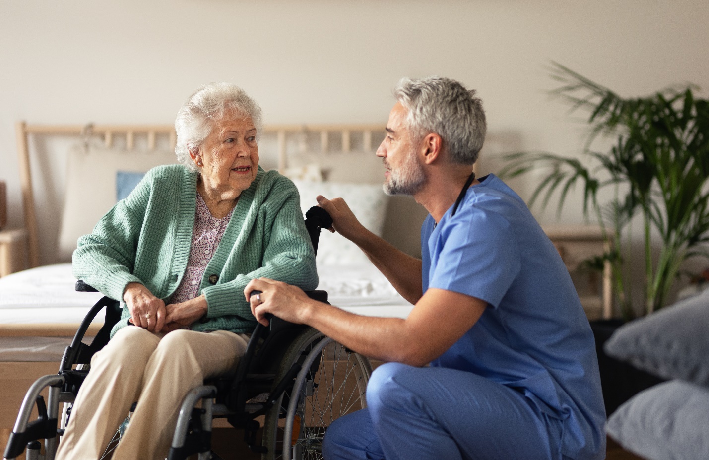 A person in a blue scrubs talking to an old person in a wheelchair

Description automatically generated