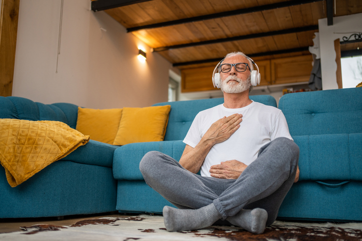 A senior man sits cross-legged on the floor, wearing headphones and practicing mindfulness with his eyes closed. | Homewatch CareGivers of Longmont