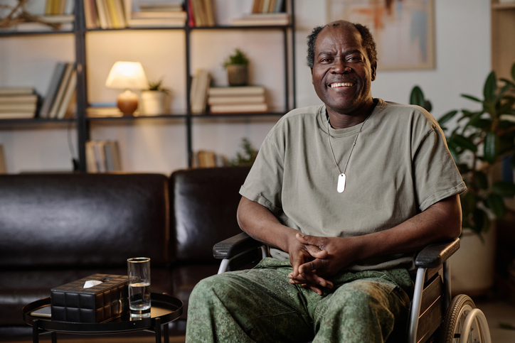 An elderly man, adorned with dog tags that signify his status as a veteran, sits in a wheelchair, smiling warmly. | Homewatch CareGivers of Arvada