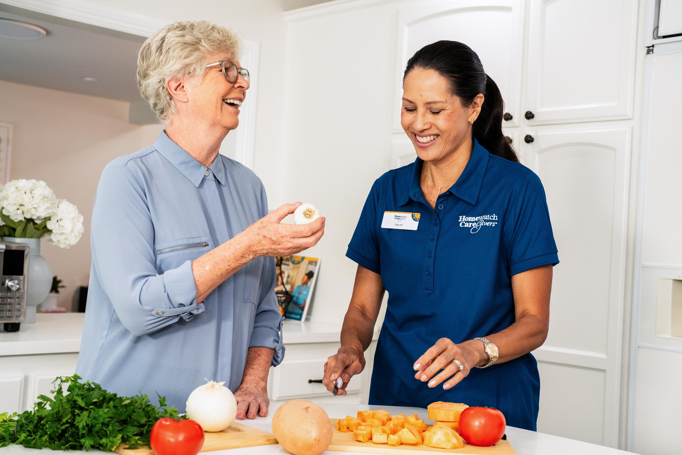 caregiver laughing with older woman in kitchen next to vegetables