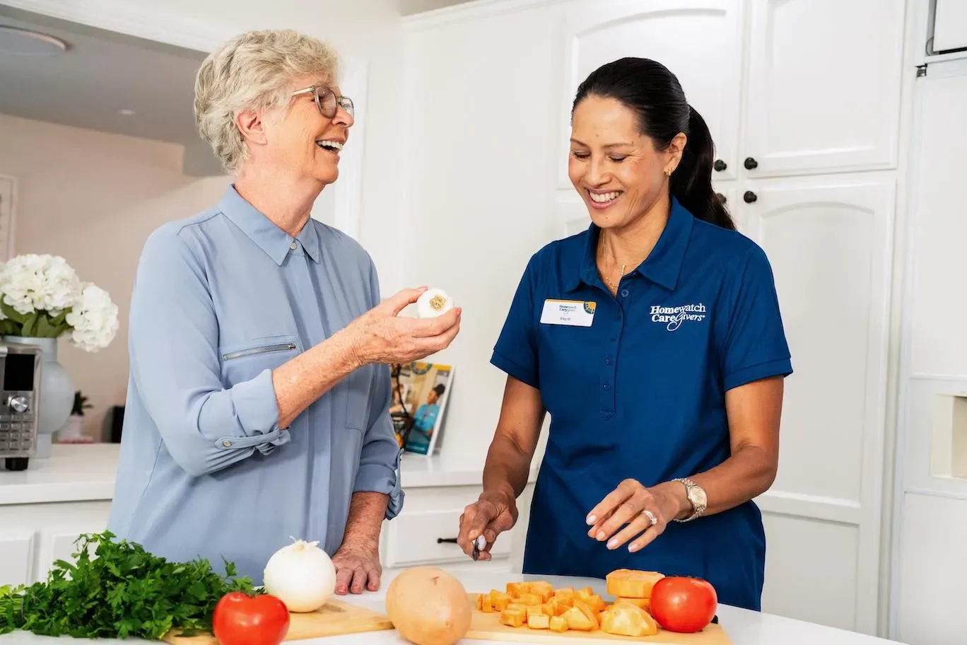 caregiver laughing with older woman in kitchen next to vegetables