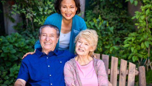 couple sitting on bench smiling with smiling caregiver behind them