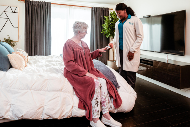 caregiver smiling and handing woman siting on bed in robe something